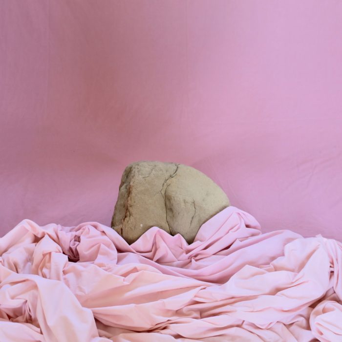 A grey rock in the shape of an arse propped up theatrically against a dusty pink wall and draped in pink sheets.