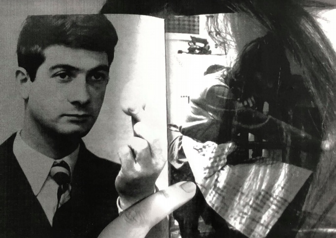 A black and white image of finger holding open a booklet showing two photographs. In the image on the left hand page of the booklet is a serious looking male presenting person holding an egg and on the right handside image is a female identifying body slumped over a table wearing an apron. The finger that is holding open the booklet is pointing to the picture of the person with the apron. 