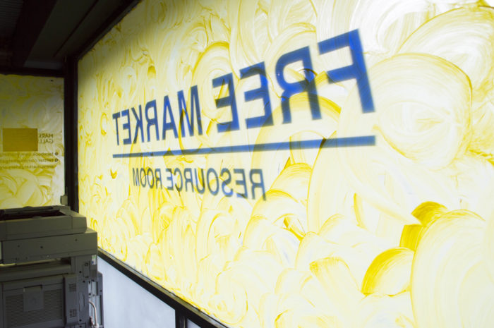 A gallery's window painted in yellow with he text Free Market Resource Room in blue capital letters reading backwards as the photograph is taken from the inside. In a corner there is a big photocopy machine. 