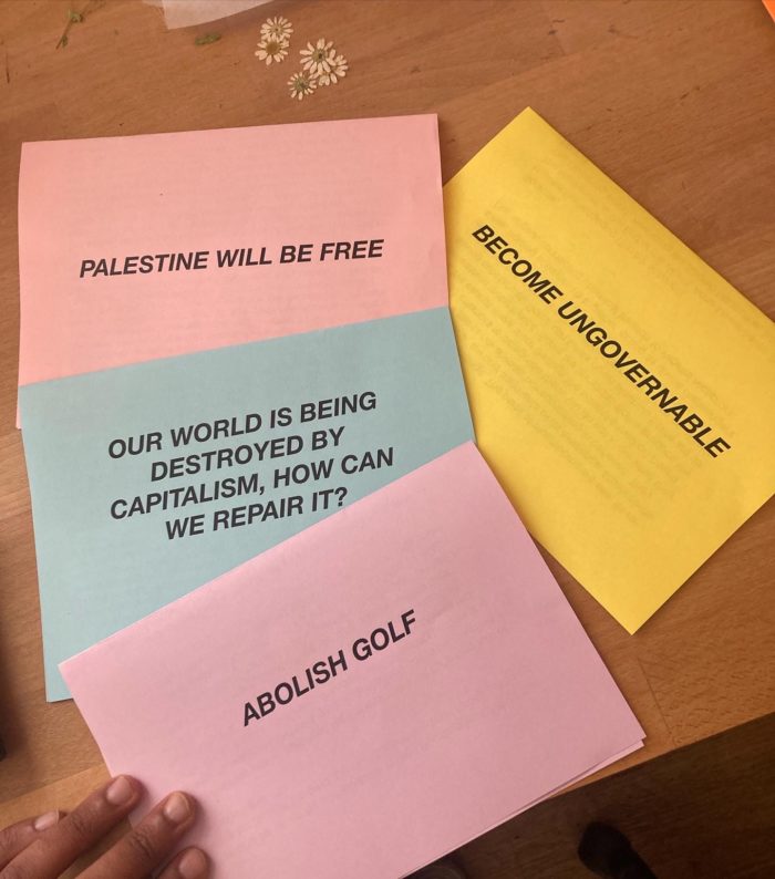 A colour image of hand of a person of colour over 4 sheets of paper in pastel pink blue and yellow reading : Abolish Golf, Our world is being destroyed by capitalism, how can we repair it?, Palestine will be free, become ungovernable. 