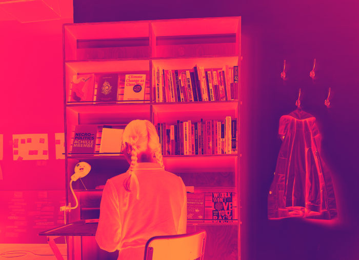 A bright pink and orange inverted image of a person with split braided pigtails seen from the back sitting in front of a bookshelf with books next to a coat hanging off a coat-hanger