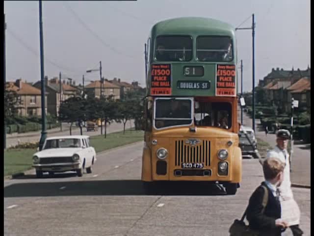 An old style white car and yellow and green bus wait on a residential street while people cross the road in front of the camera.