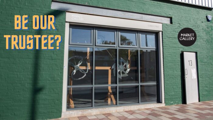 a poster with the bold yellow text 'be our trustee?' at the bottom, and the market gallery logo, in a black circle at the top. The background is a photo of the gallery outside, a shop front with green brick and four big windows, and a sculpture inside made of a wooden frame and metal wheels.