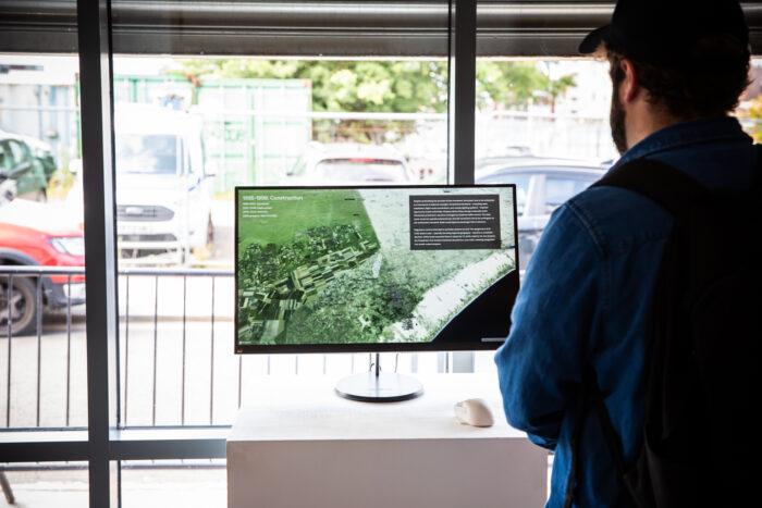 A person wearing a cup looking at a monitor with an aerial photograph. Behind the monitor, the street with cars can be seen.