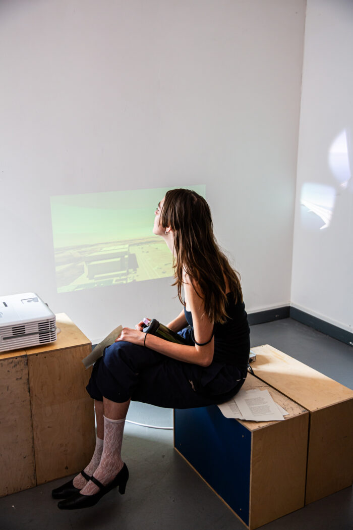 A photo of a person with long hair and white laze shock with hills sitting on a wooden box in front of a projection.