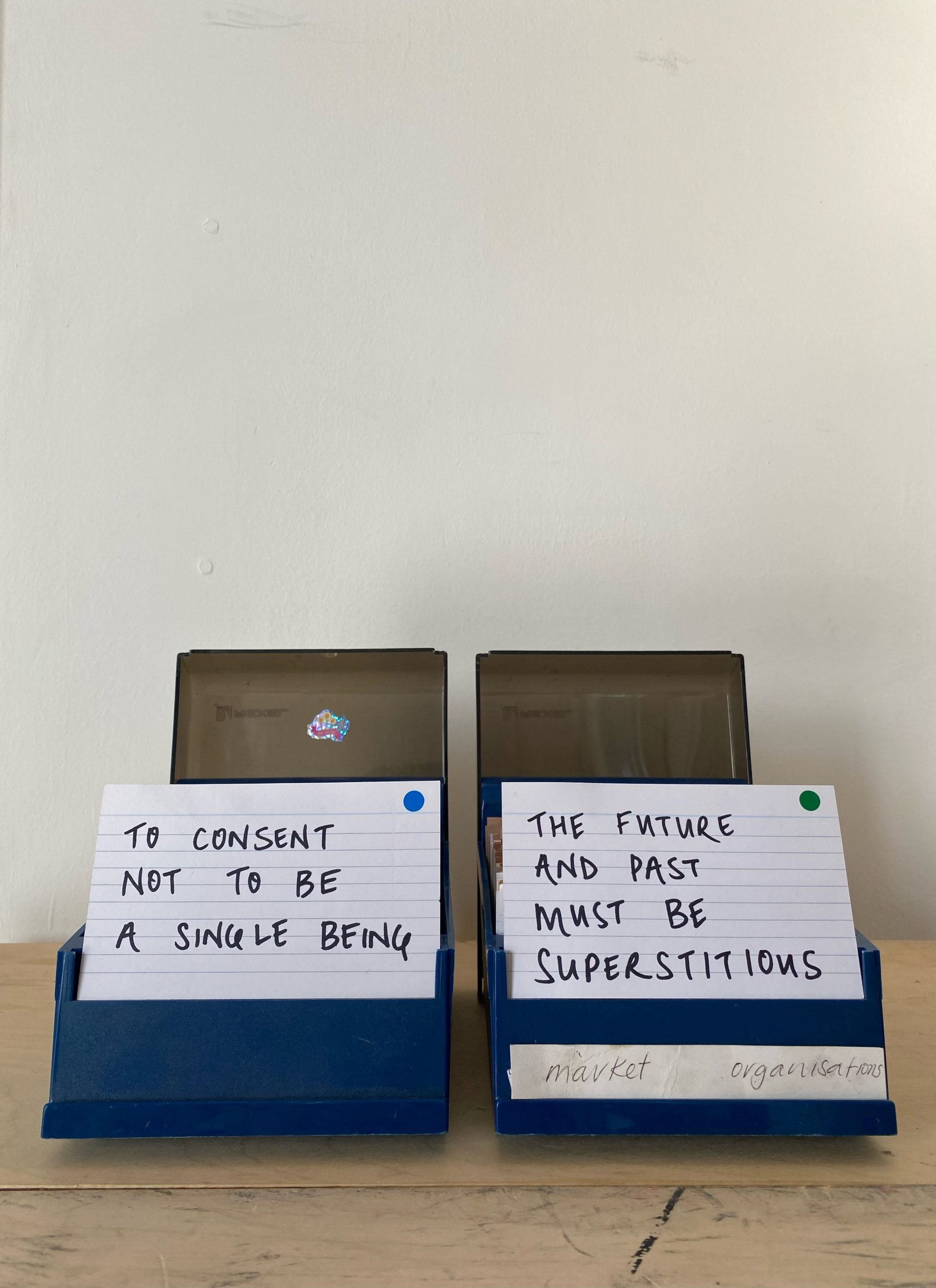 Two blue plastic card holders sitting side by side against a white wall on a wooden surface with two cards reading 'to consent not to be a single being' & 'the future and past must be superstitious'