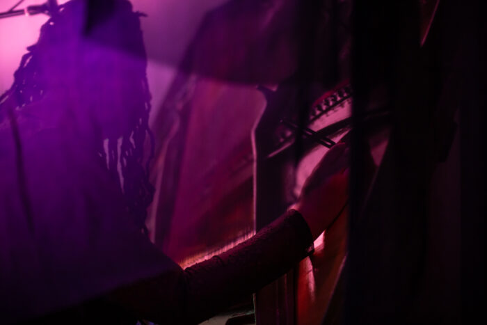 A close up of Ruby Allen positioned a tuner in the piano harp. She is sitting behind a veil of blue gauze that is light up in pink and purple colours