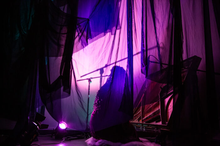 Ruby Allen kneeling on the floor in front of the piano harp with a microphone stand next to her. She is sitting behind a veil of blue gauze that is light up in pink and purple colours