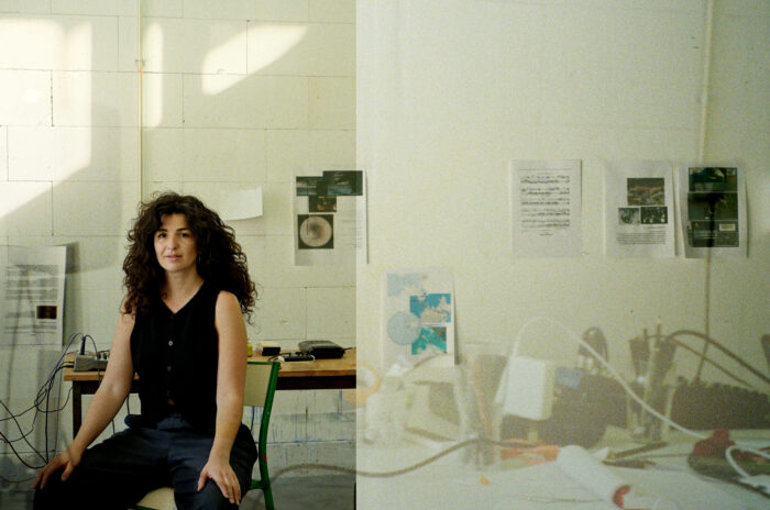 Artist Hannan Jones sitting on a chair in a studio with her hands resting on her thighs. She has long curly hair and is wearing a black vest. 