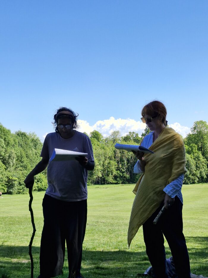 Two people standing under the shade of a tree reading from a sheet of paper in a park with trees on a sunny day.