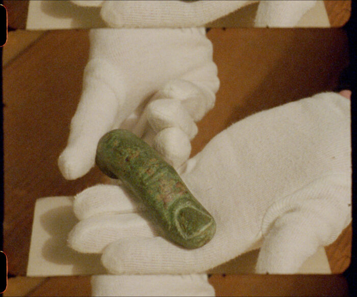 Hands in white gloves hold a single finger carved out of green stone against a warm brown background. The finger sits large in the hands, like that of a giant.