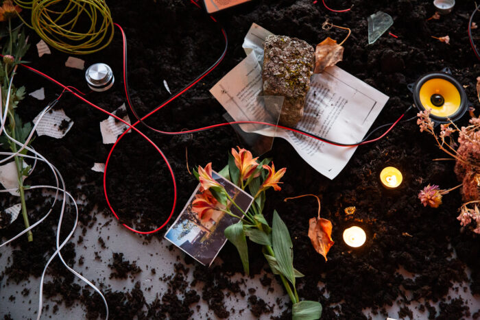 A close up of an installation with soil flowers, rocks, cables, candles and speakers.