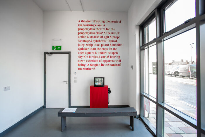 An old crt tv playing a film over a plinth covered with red fabric. On the right side of the image there is a big street facing window and behind the tv a handwritten mural with red letters. 