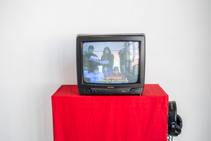 A close up of a crt tv playing a film with 6 people reading a play. The tv is standing on a plinth covered with red fabric and on the right side of the plinth a pair of headphones are hanging. 