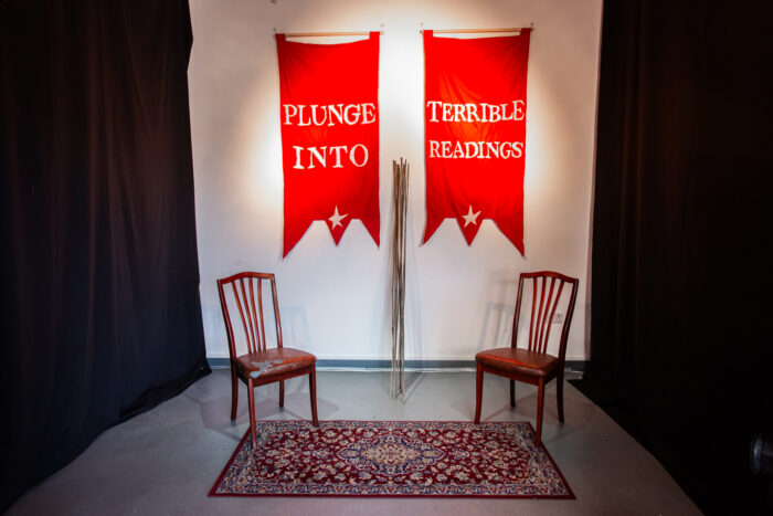 Two red fabric banners hung up on a white wall with the words 'plunge into' and 'terrible readings' written in white. In between the banners there is a bundle of canes and just in front there are two old chairs placed on top of a Persian style carpet. 