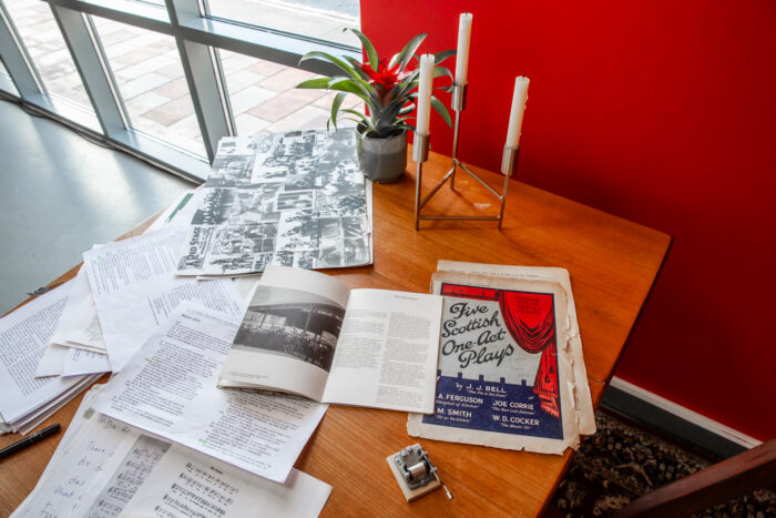 A wooden table with several old sheets of paper laid out as well as a golden candle holder and a potted plant. The table is next to a red wall. 