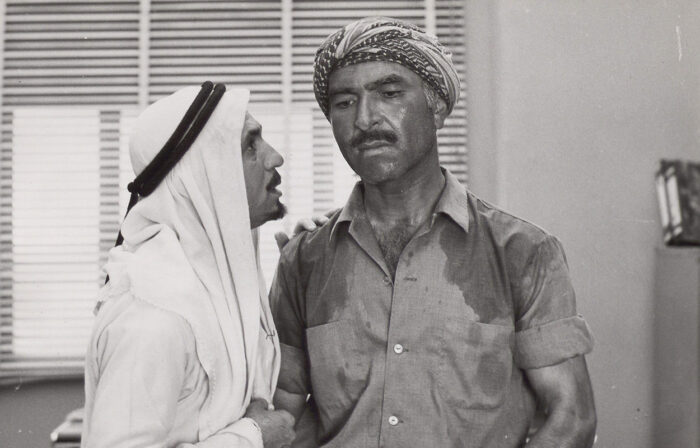 Two people on a room, the one on the left wearing a white shemagh headscarf with an agal is holding the  the right person's shoulder who is wearing a Keffiyeh wrapped around his head and a shirt that has sweat patches. The face of the person on the right is pensive and concerned. 