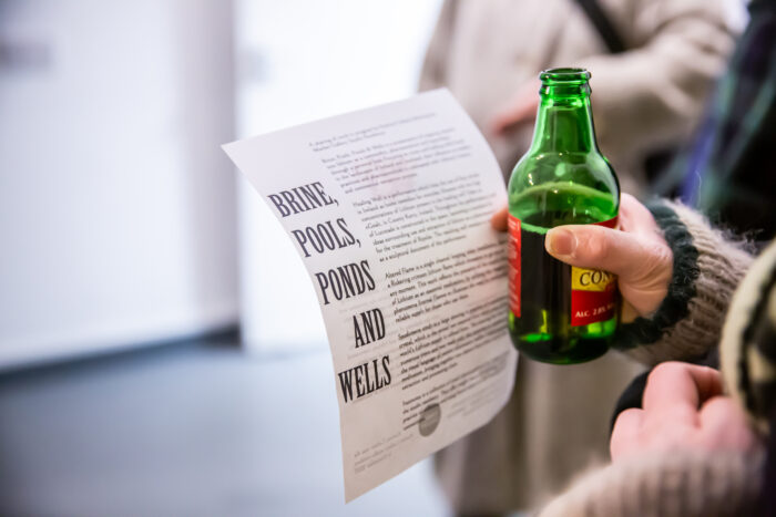 A hand holds a bottle of beer and a paper flyer. The text BRINE POOLS PONDS AND WELLS is on the right of the paper with a body of text on the left out of foucs
