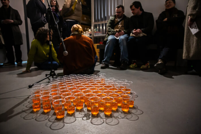 A group of plastic cups, half filled with an orange liquid are on the floor. A mic points towards them and their is a group of people sitting in the background out of focus.