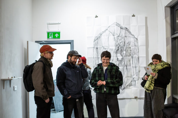 Image of the Artist among peers, smiling within Gallery space. Behind the group we can see a large pencil drawing, which has crease lines across it.