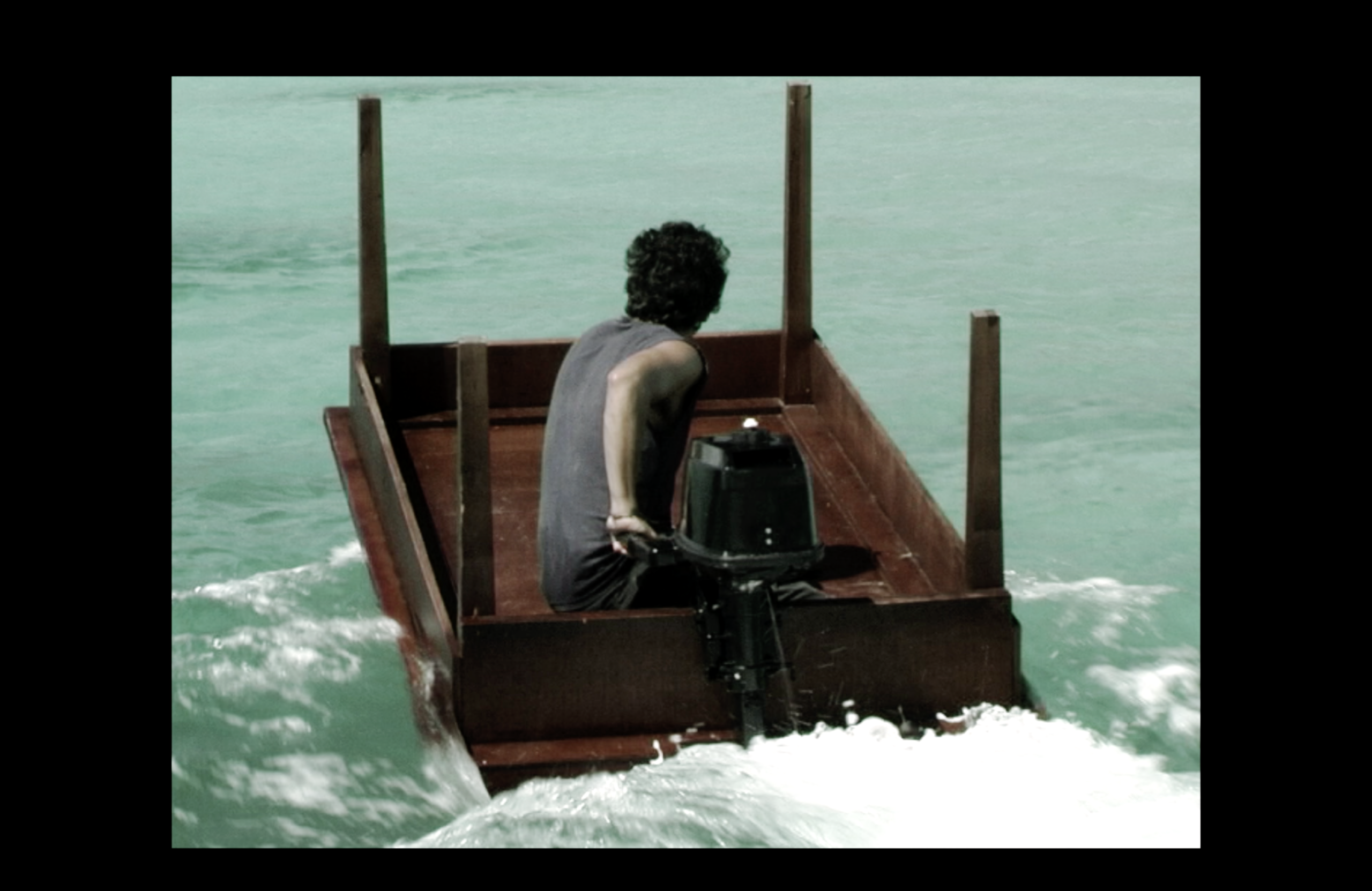 A grainy colour image shows a person seated inside an upside-down table with a black outboard motor attached. The makeshift vessel cuts through pale turquoise water, churning the surface behind it, creating white foam. The figure is turned away from the camera, their bare arm catching the light, hand on the tiller.