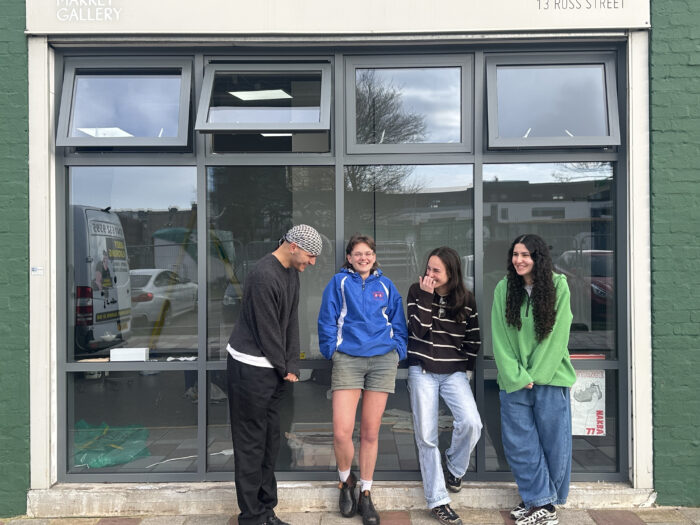 Four people standing outside a large window in front of an art gallery. At the top of the window there is a panel that reads "Market Gallery" on the left and "13 Ross Street" on the right. The four people are mid-laughing and standing close together.