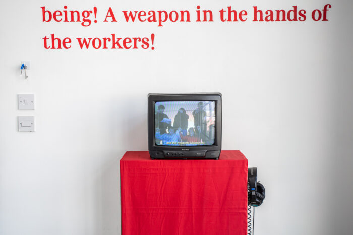 An old crt tv playing a film over a plinth covered with red fabric with headphones hanging on the right hand side. Above the tv there is red writing on the wall in large font that reads "being! A weapon in the hands of the workers!"