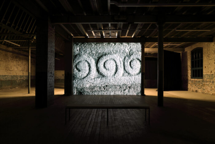 A dark exhibition space with a large film projection in the centre with a backlight. The image being projected is a close up of a rock's surface with three swirling engravings. 