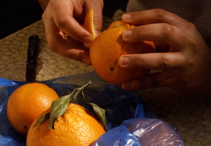 Close up shot of a pair of hands peeling an orange next to two other oranges that are yet to be peeled. They are cradled in a blue plastic grocery bag. The handle of a knife can be seen nearby on the table that everything is resting on. The still is from footage shot on 16mm.