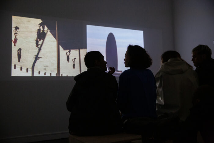 The silhouette of four people sitting on a bench in a dark gallery space with their back to the camera. They are watching a two-channel moving image work which is playing in the background. The left channel of the work shows video taken of a mirrored surface reflecting seven people walking on the street below. The right channel of the work shows a thin oval silhouette against a hazy sky.