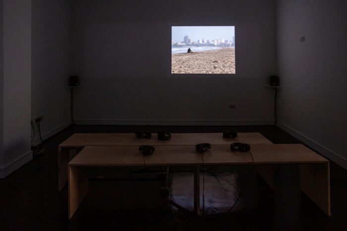 A photo of a projected video work on the right hand side of a wall in a dark gallery space. The video shows a person sitting down on the sand at the beach, with the sea and a skyline of buildings in the background. In the foreground of the image there are two benches back to back with six sets of headphones resting on top.