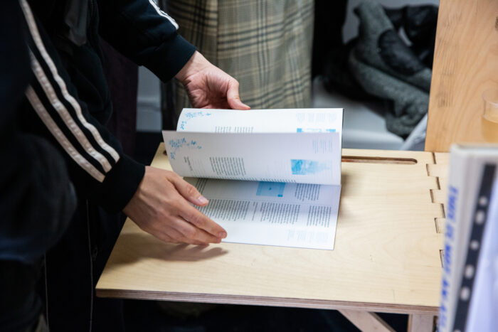 A photograph of a person flicking through a publication resting on a wooden display shelf. The publication is on white paper with blue images and dark green text which is not visible or legible in the image. The person is wearing a long sleeve black adidas top and only their arms are visible in the photograph.