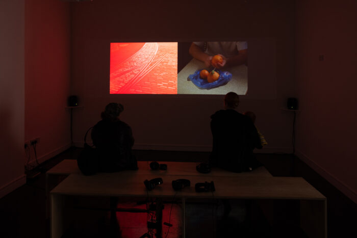 A two-channel video work projected on to a wall in a dark gallery space. Almost non-visible, two people sit on the far end of a bench facing the work. The left channel shows tram lines on a cobbled road with a red haze over the top. The right channel shows a person peeling an orange with two other oranges nestled in a blue plastic bag close by. The image is close up so you can only see the person's arms and the oranges, bag and table top.