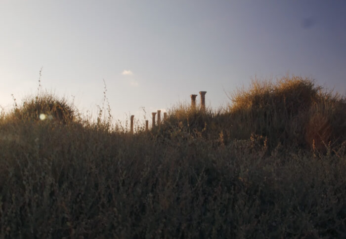Seven stone columns standing at varying heights can be seen in the distance, behind a grassy hill in the foreground. The footage could have been taken in the early hours of the morning or at dusk. The sky is a very pale powder blue with one tiny, cloud in it.