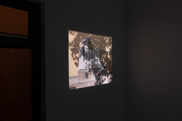 A projected image in a dark space showing a statue of a person slightly reclined with one arm hanging down to their side and the other with their head resting on top. The statue is raised from the ground and sits in front of a yellow wall. The shadow of a tree covers the woman with dappled sunlight coming through. The video has been taken outdoors.