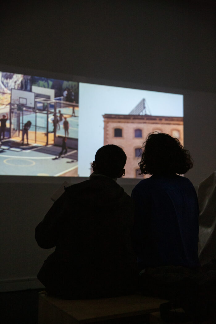 The silhouette of two people sitting on a bench watching a two-channel moving image projected on to the wall in a dark gallery space. The left channel of the video work shows a group of people playing basketball outdoors in the sun. The right channel shows a building with boarded up windows.
