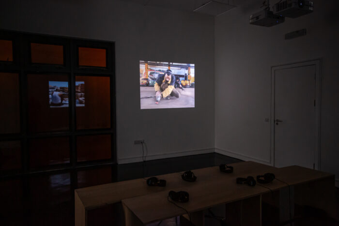 A photo of a projected video work in the corner of a dark gallery space. The video shows a person knelt down sanding a concrete floor. In the background there are unused bumper cars. There is a reflection in the window in the space to the left of the projected image which faintly shows an image of a person sitting down at a dock overlooking a block of flats. In the foreground of the image there are two benches back to back with six sets of headphones resting on top.