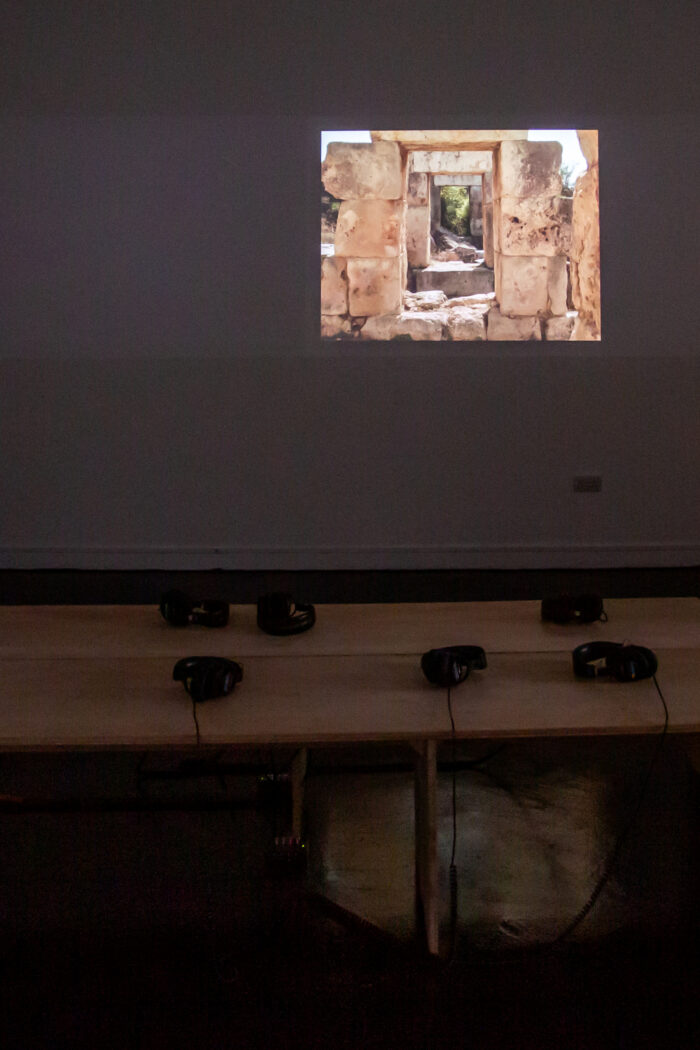 Photograph taken in a dark gallery space with a projected image on the right side of the wall, showing a series of sandstone archways made of large rectangular stones. In the foreground of the photograph, there are two benches sitting back to back with six sets of headphones visible.