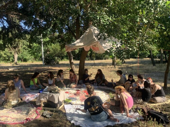 Documentation of Frieda Ford's workshop that took place in Athens, Greece: a group of people sit on a green populated with trees. The group are sitting in a circle having conversations in pairs, atop a patchwork of blankets and sitting off-centre is a small table with a pile of folders on top. Above them hangs from a tree a neutral tone and peach coloured canopy. The sun shines dappled across the dry, sun-bleached grass landing on some of the group, whilst others are sheltered in the shade.