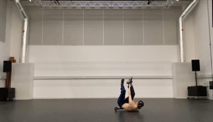 The artist falls onto their back in a large studio, balancing a micro-scooter between raised legs.