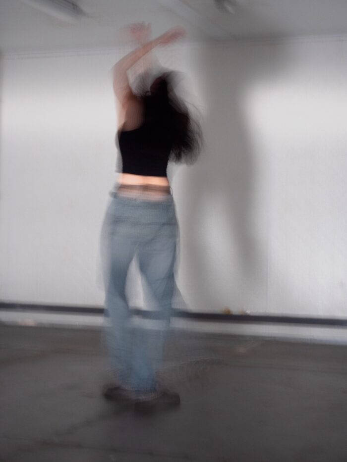 A person with long dark hair, wearing blue jeans, a black short-sleeved top and black shoes, photographed mid-movement with their arms in the air. Their image has been captured with their back facing the camera. They are in a white-walled room with a blue/black gloss floor. The entire image is out of focus.